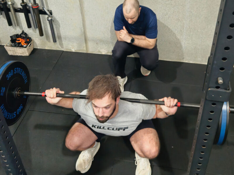 A client performing a barbell squat while a physiotherapist observes.