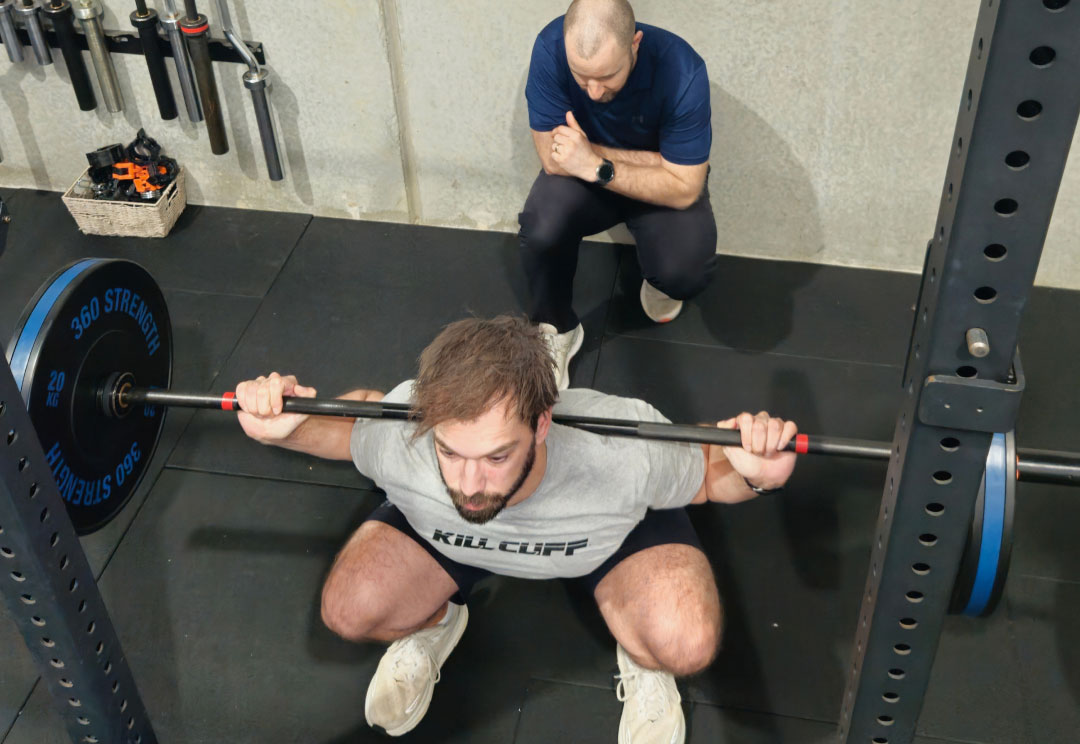A client performing a barbell squat while a physiotherapist observes.