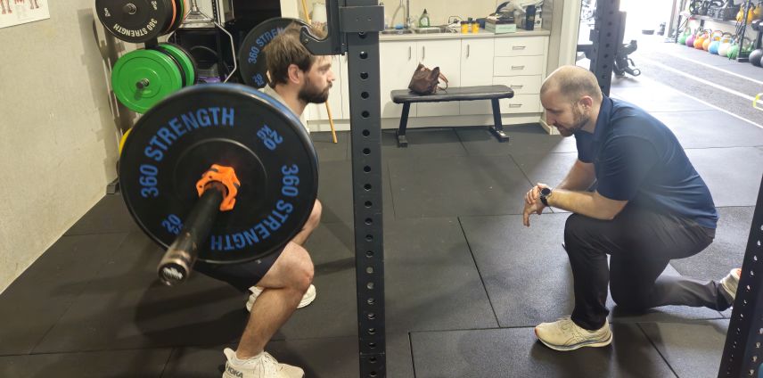 Physiotherapist observing an athlete performing a barbell back squat in a gym rig