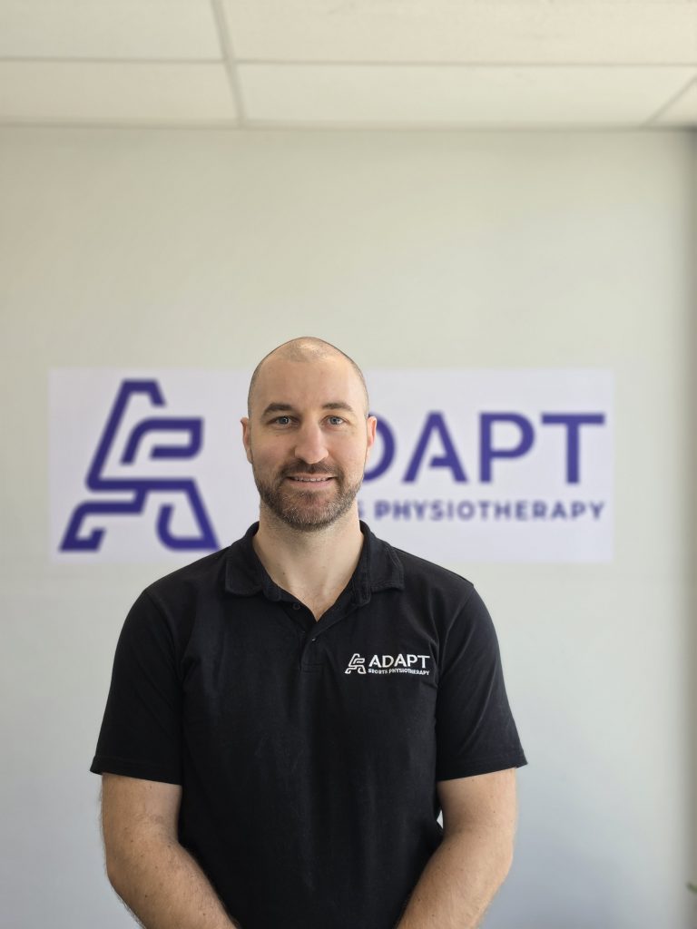Ryan Harris, physiotherapist at Adapt Sports Physiotherapy, standing in front of the Adapt signage.