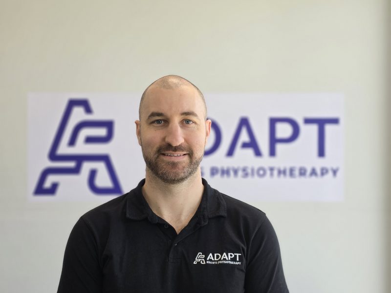 Ryan Harris, physiotherapist at Adapt Sports Physiotherapy, standing in front of the Adapt signage.